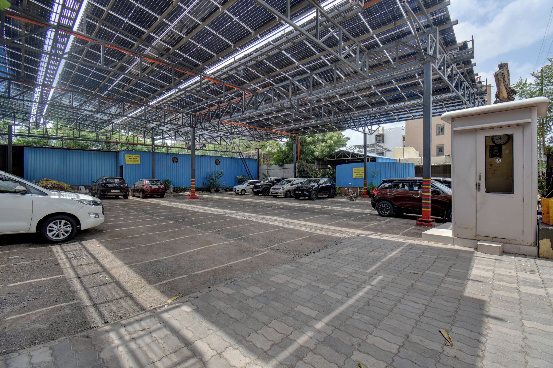 Un estacionamiento con varios automóviles, una estructura cubierta con paneles solares y un pequeño edificio de servicios.