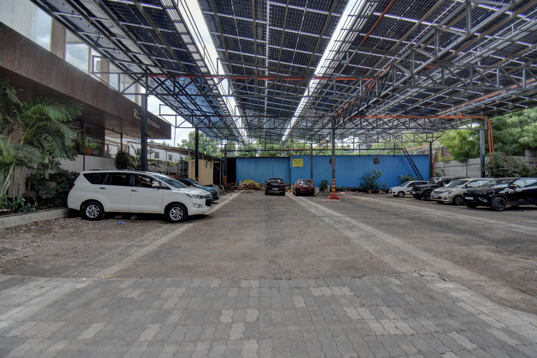 Un espace de stationnement couvert où sont garées plusieurs voitures, un conteneur bleu et un bâtiment doté d’une structure de toit en métal.