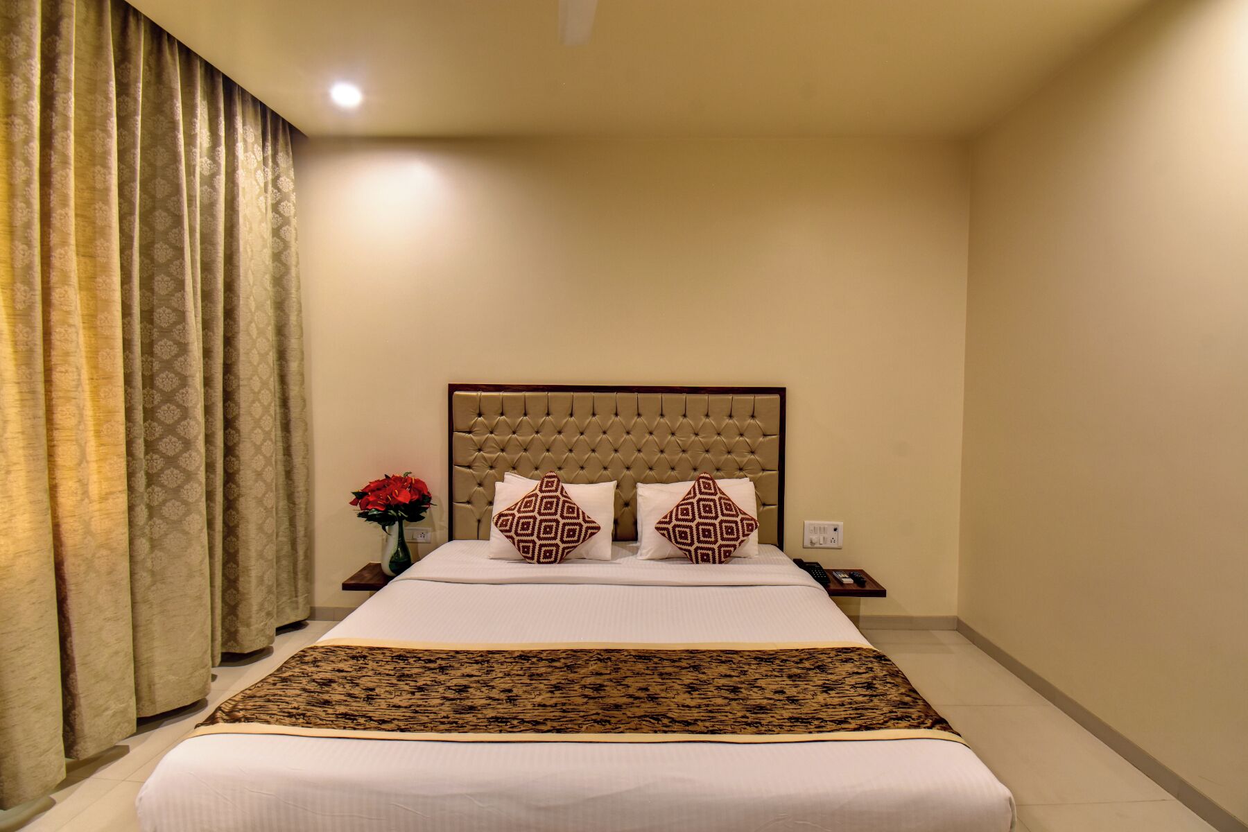 Une chambre d’hôtel avec un lit, une table de chevet, un vase avec des fleurs et un rideau.