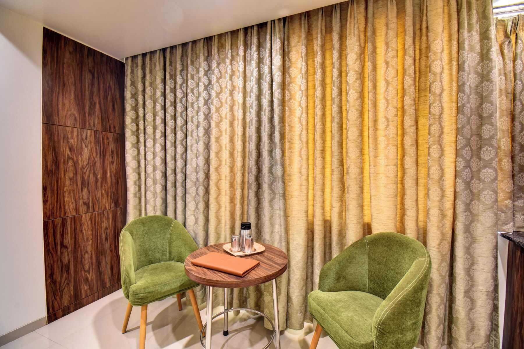 A room with two green armchairs, a small wooden table with a laptop and a thermos, and a patterned curtain backdrop.