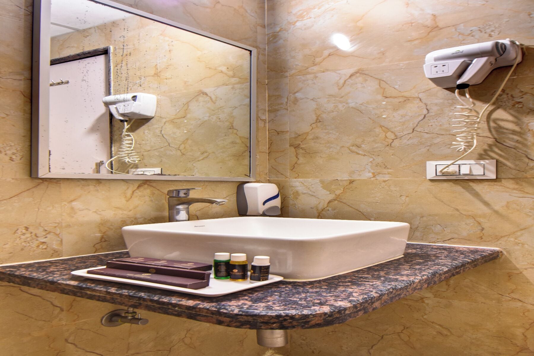 Bagno con parete in marmo, lavandino con rubinetto, specchio, asciugacapelli e un vassoio con articoli da toilette.