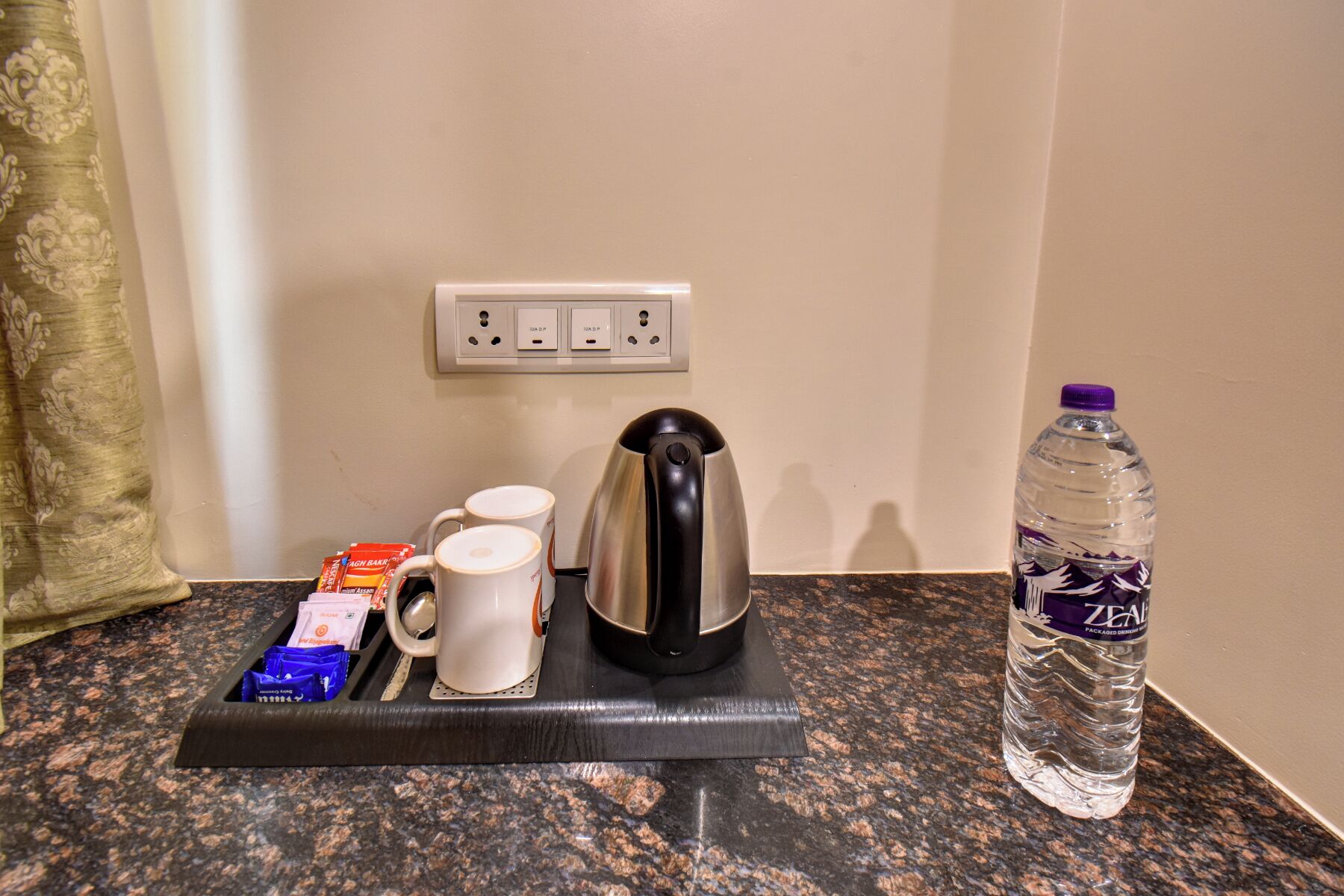 A kitchen counter with a kettle, two mugs, a water bottle, and some packaged snacks.