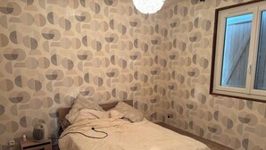 Um quarto com papel de parede estampado, uma cama de madeira, uma mesa de cabeceira com um ventilador e uma janela com persianas.