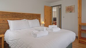 Una cama bien hecha con sábanas blancas y toallas enrolladas, cabecera de madera, mesita de noche y televisor montado en la pared.