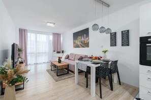 Moderni olohuone, jossa on sohva, sohvapöytä, ruokapöytä ja tuoleja. Seinällä on televisio, kasvi ja kaupunkimaisemaa esittävä taulu.
