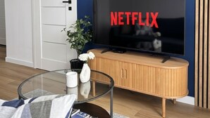A modern living room with a flat-screen TV, a wooden cabinet, a glass coffee table, and a potted plant. - Lumina Comfort Apart near ZOO Orientarium with Parking (Łódź)