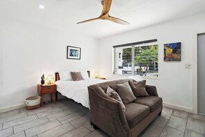 Ein modernes Schlafzimmer mit Bett, braunem Sofa, Nachttisch aus Holz, Stehlampe und Deckenventilator.