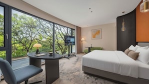 Habitación de hotel moderna con una cama grande, un escritorio con lámpara, televisión y vistas a árboles a través de amplios ventanales.
