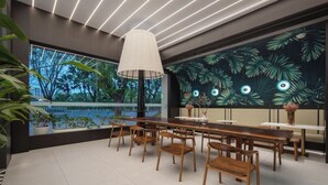 Un comedor moderno con mesas y sillas de madera, un ventanal con vistas a árboles y un mural en la pared con hojas tropicales.