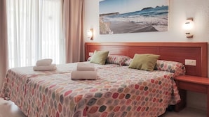 Quarto de hotel com cama, mesa de cabeceira, quadro de uma praia e janela com cortinas.