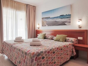 Ein Hotelzimmer mit Bett, Nachttisch, einem Strandbild und einem Fenster mit Vorhängen.