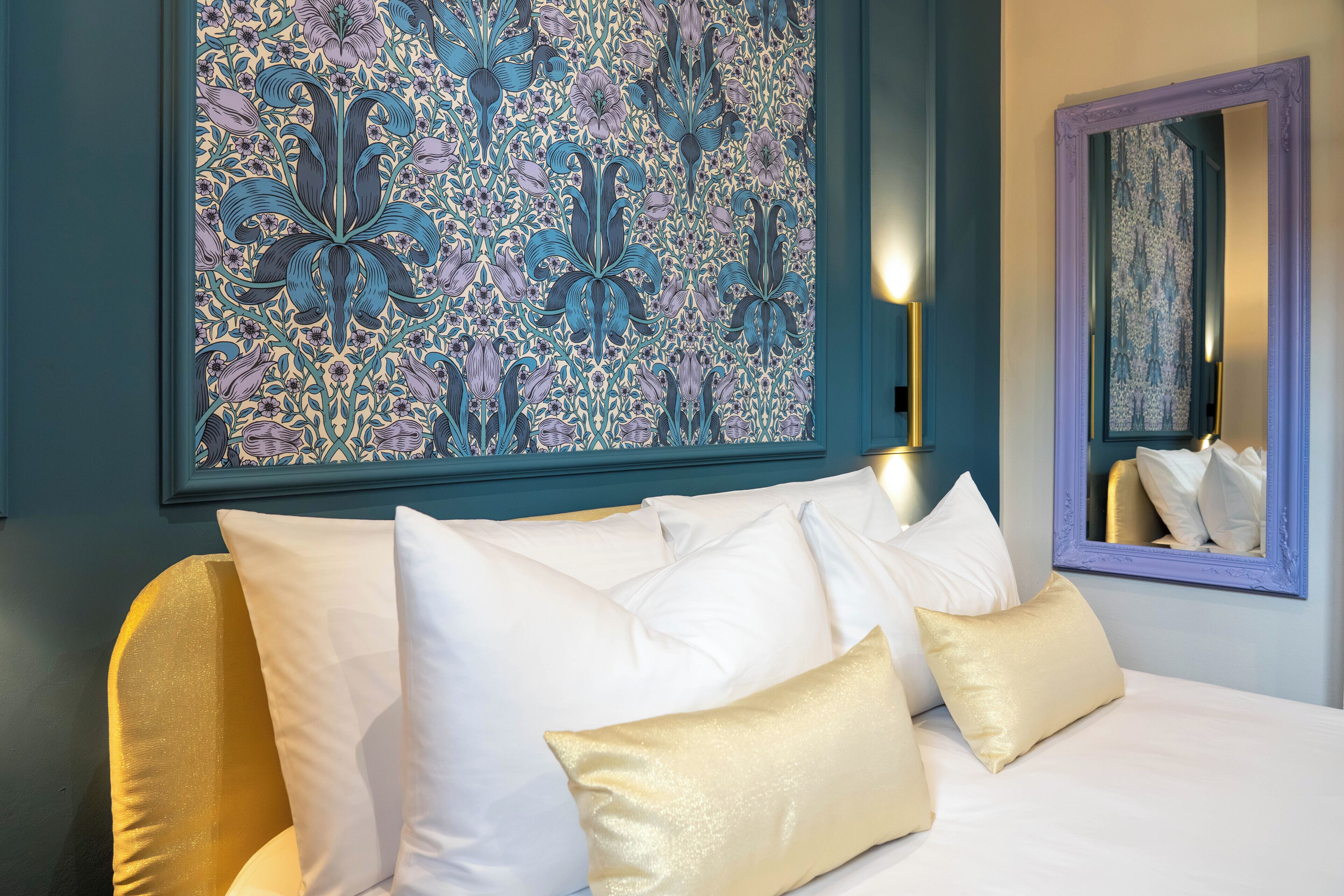 Een bed met witte en gouden kussens, een groot kunstwerk met bloemenprint en een spiegel met paars frame.