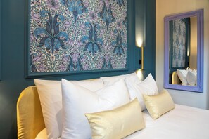 Un lit avec des oreillers blancs et dorés, une grande œuvre d’art à motifs floraux et un miroir avec un cadre violet.
