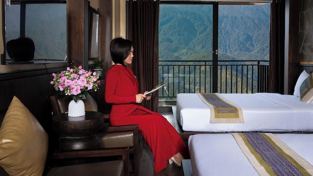 Quarto de hotel com cama, poltrona, mesa pequena com vaso de flores e vista para montanhas.