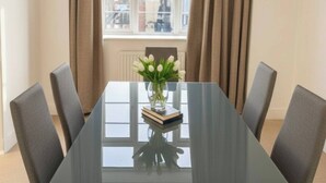 Un comedor moderno con una mesa de vidrio, sillas grises, un florero con flores blancas y una ventana con cortinas.