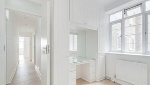 Una habitación luminosa y blanca con un escritorio de vidrio, armarios blancos y un ventanal grande.