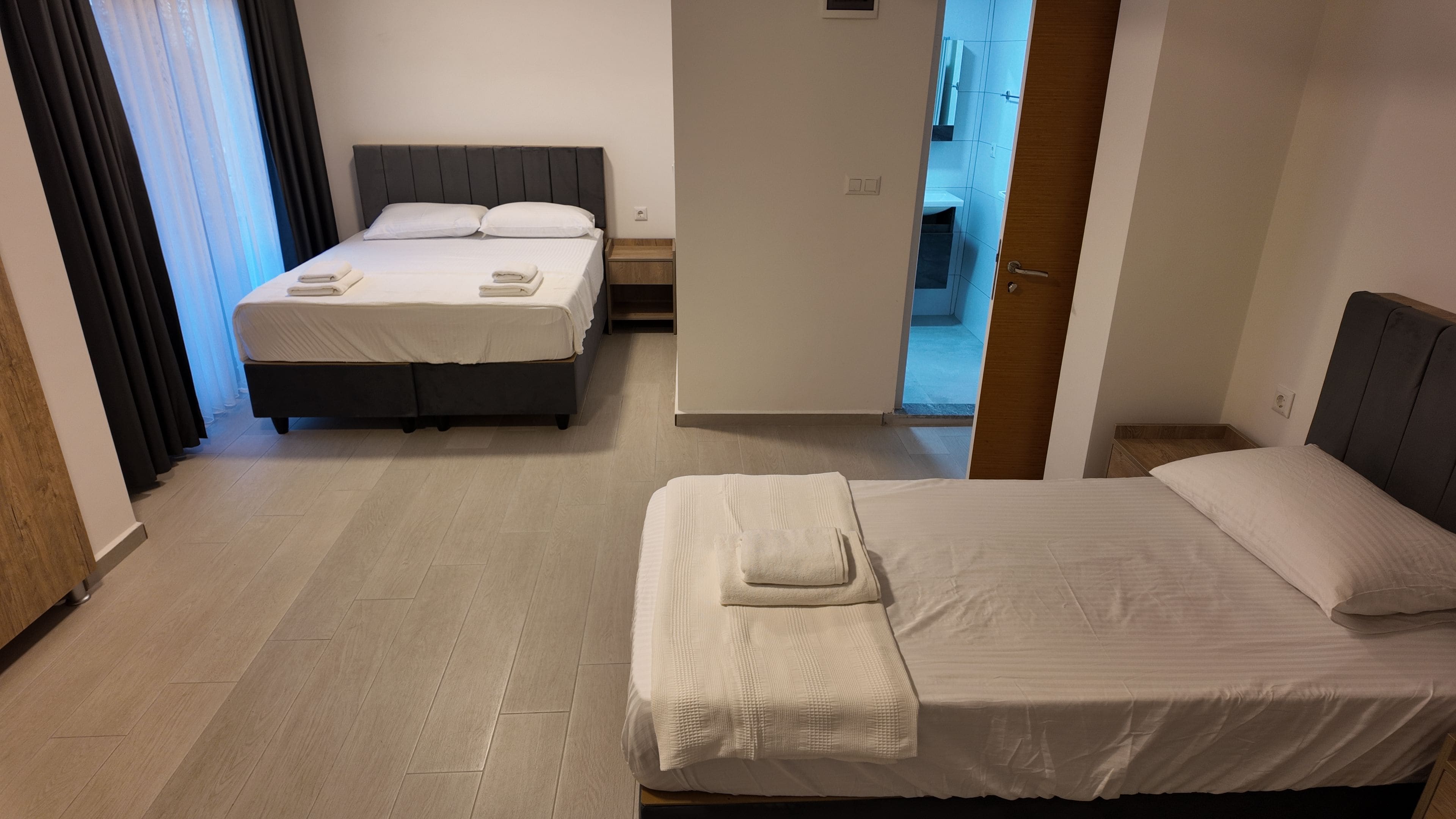 Hotellihuone, jossa on kaksi sänkyä, puulattia, ovi, joka johtaa kylpyhuoneeseen, ja ikkunasta avautuva näkymä ulos.
