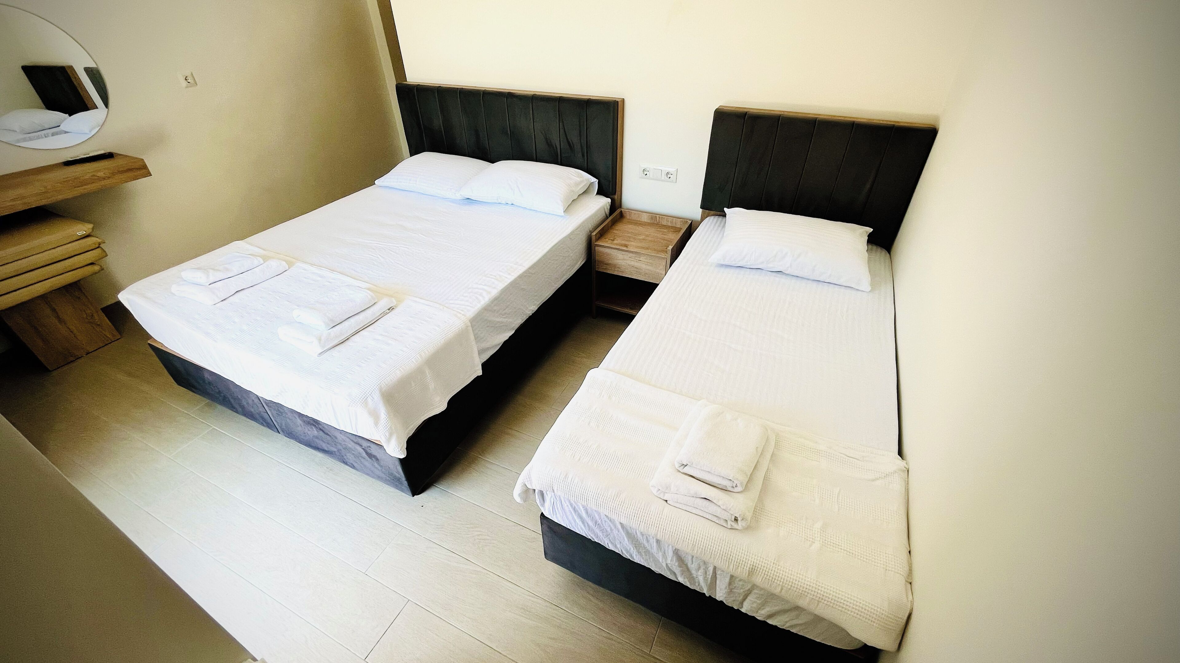 Dos camas individuales con ropa de cama blanca, un banco de madera, una mesita de noche de madera y un espejo fijado a la pared.
