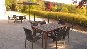 Een terras buiten met een houten tafel en stoelen, omringd door een metalen hek, met uitzicht op een aangelegde tuin.