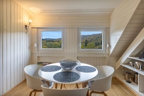 Comedor con una mesa redonda, cuatro sillas y dos ventanas que dan a una ladera boscosa.