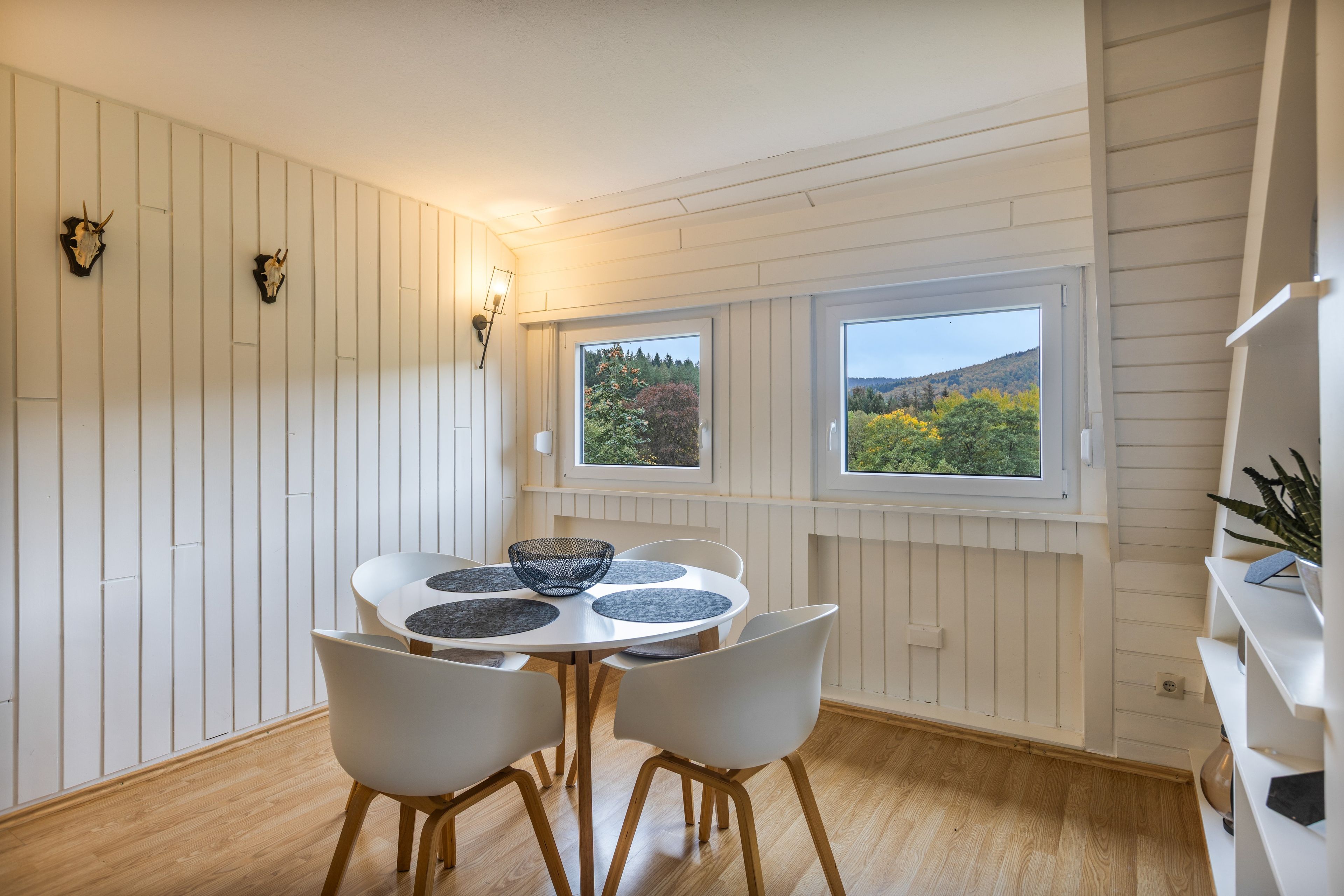 Un comedor con una mesa redonda, cuatro sillas y dos ventanas que dan a un paisaje boscoso.