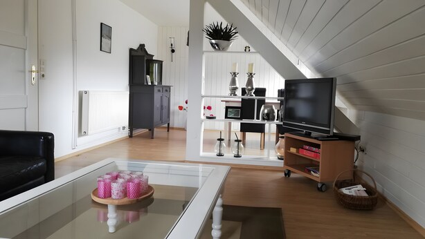Ett vardagsrum med ett soffbord i glas, en svart soffa, en tv på ett tv-bord, en bokhylla och en trappa.