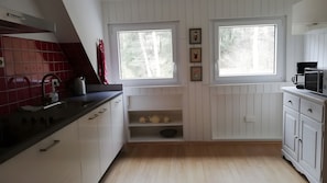 Eine Küche mit weißen Schränken, einer schwarzen Arbeitsplatte, einem Wasserkocher und einer Kaffeemaschine. Zwei Fenster bieten Blick auf Bäume.