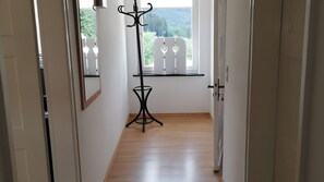 Ein Flur mit Holzboden, einer schwarzen Stehlampe, einem Spiegel und einem Fenster mit Blick auf Grünflächen.