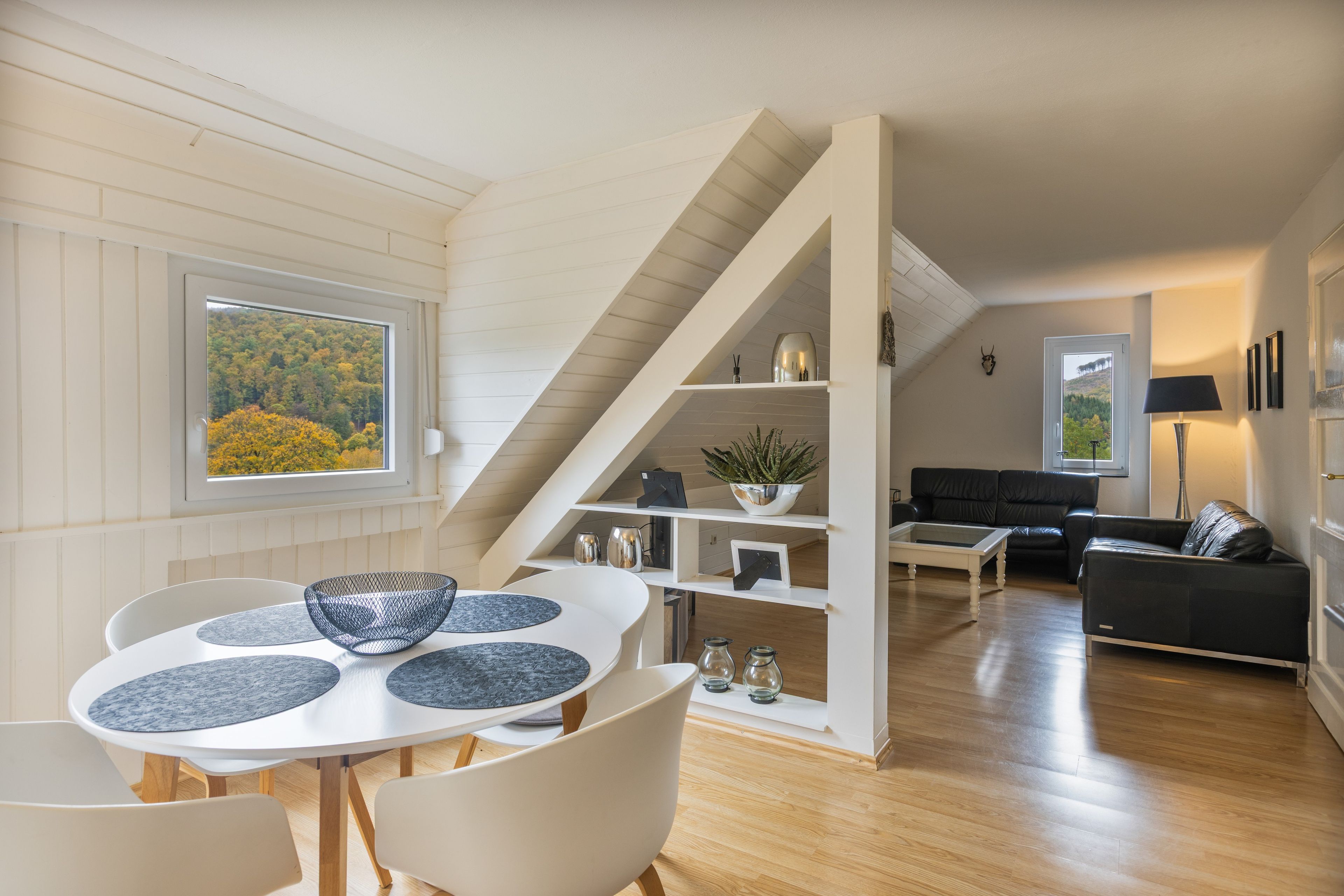 Ein modernes Wohnzimmer mit Treppe, einem Essbereich mit weißem Tisch und Stühlen sowie einem schwarzen Sofa.