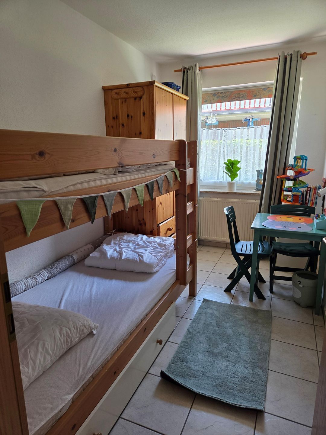 A bunk bed with a wooden headboard, a desk with a chair, a radiator, and a window with curtains.
