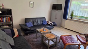 A living room with a black leather sofa, a grey cabinet, a television, and a window with curtains. - Vacation Rental at Am Harlebogen 11, Apartment 1 - Vacation Rental at Am Harlebogen 9, Apartment 1 (Wittmund)