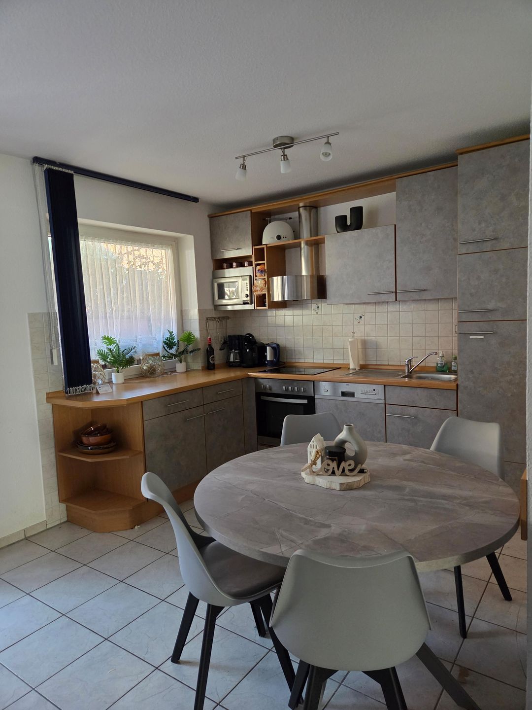 Una cocina moderna con comedor, que incluye una mesa redonda con sillas, electrodomésticos integrados y una ventana con cortinas translúcidas.
