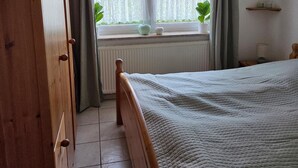 A bedroom with a bed, wooden wardrobe, window with curtains, radiator, and a shelf with items. - Vacation Rental at Am Harlebogen 11, Apartment 1 - Vacation Rental at Am Harlebogen 9, Apartment 1 (Wittmund)