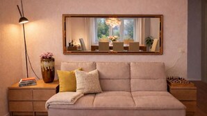 A neutral-toned sofa with a cushion, a side table with a lamp, a vase, and books. A large mirror hangs above a window with a view of greenery.