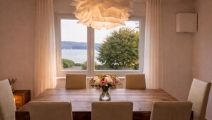 A dining area with a wooden table, four chairs, a vase of flowers, and a view of a lake through large windows.