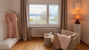 A room with a large window, a radiator, a chair, a bedside table, and a hanging robe.