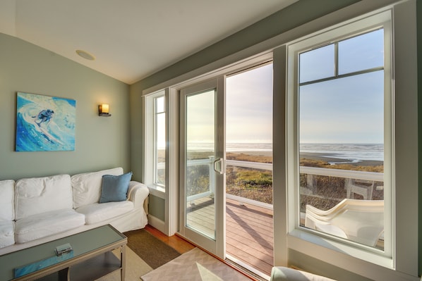Un salon avec un canapé blanc, une table basse en verre et un tableau accroché au mur. De grandes fenêtres offrent une vue sur la plage.