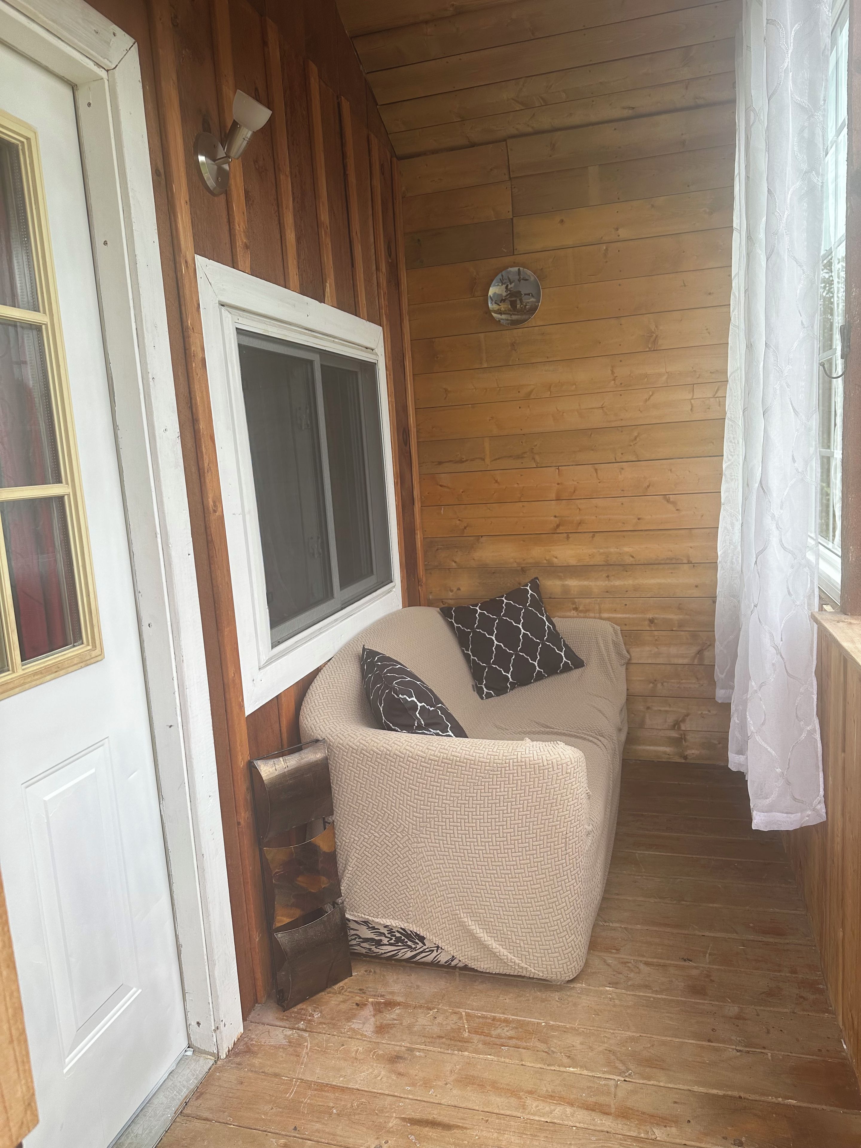 A cozy room with a wooden wall, a white door, a window with a white curtain, and a beige sofa with patterned cushions.