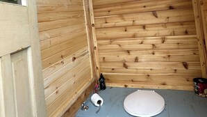 A wooden cabin interior with a single toilet, a window, and a small shelf with items.
