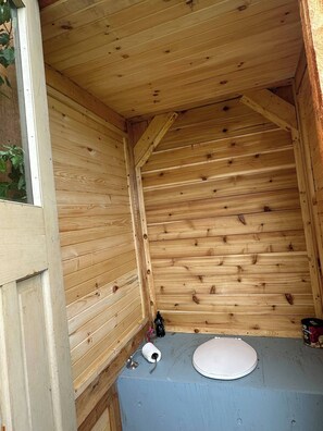 A wooden cabin interior with a single toilet, a window, and a small shelf with items.