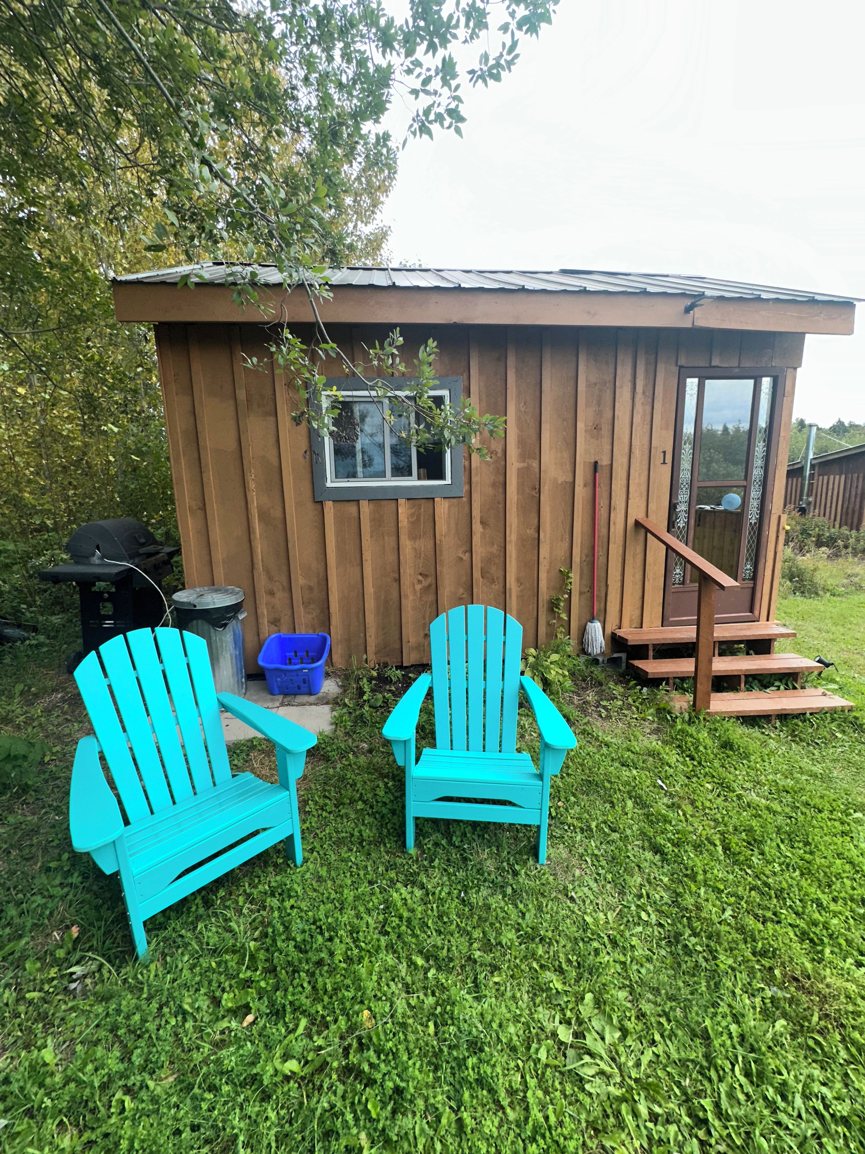 A wooden cabin with a small porch, two turquoise Adirondack chairs, and a window overlooking the yard.