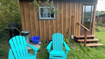 A wooden cabin with a small porch, two turquoise Adirondack chairs, and a window overlooking the yard.