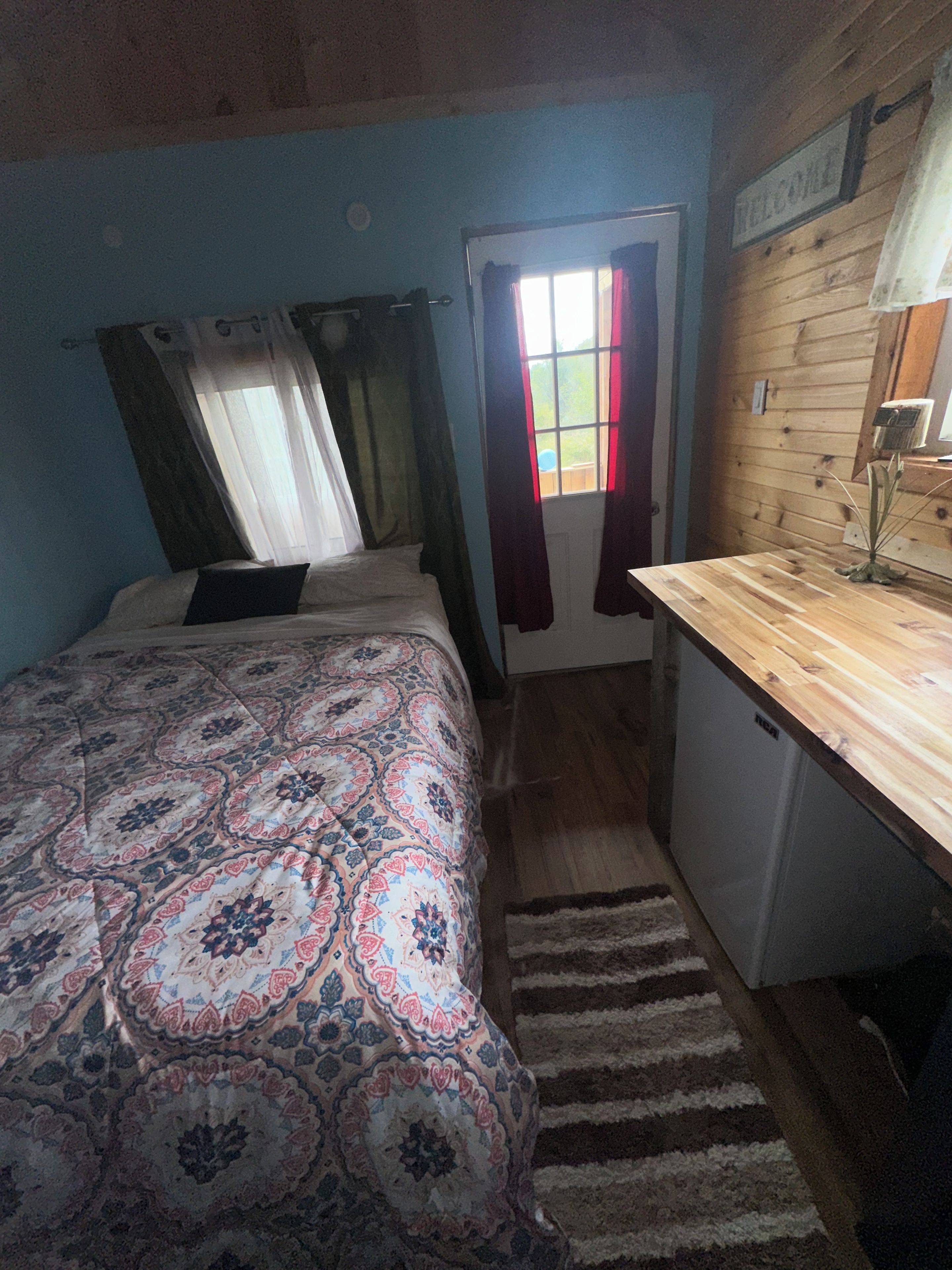 A bedroom with a bed, a desk, a window with curtains, and a door.