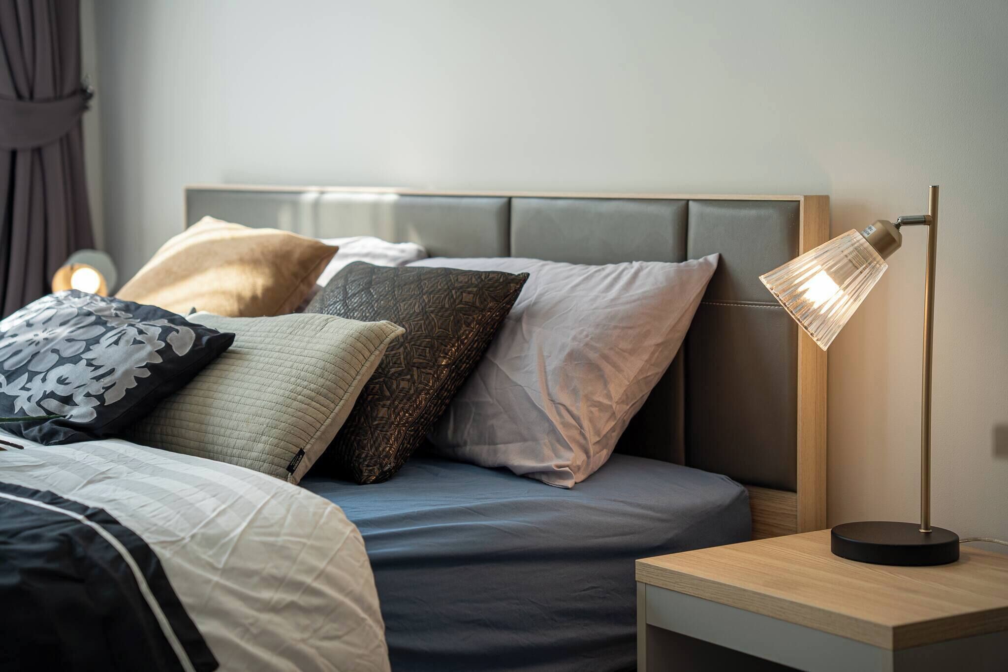 Una cama bien hecha con cabecera, una lámpara de noche y una mesita de noche de madera.