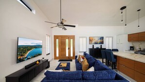 A modern living room with a large flat-screen TV, a blue sofa, a ceiling fan, and a kitchen area with wooden cabinets and a marble countertop. - Luxe Villa in the Caribbean • Gated • Pool & Sun (Sosúa)