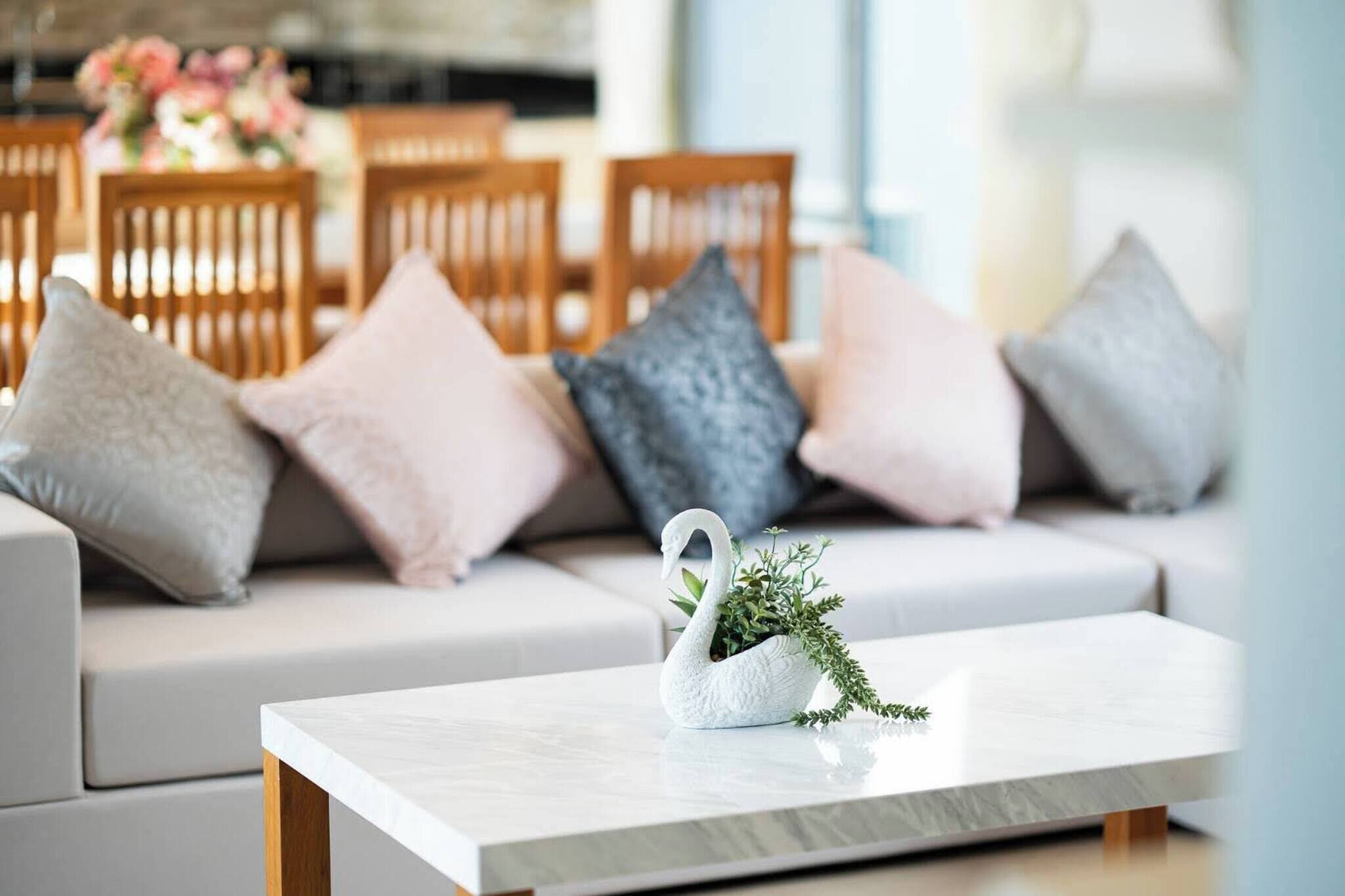 Una sala de estar moderna con un sofá blanco, cojines grises y rosas, un florero con forma de cisne blanco y follaje, y una mesa de centro de madera.