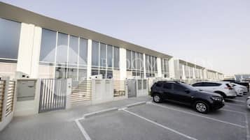 Um edifício moderno com grandes janelas, um estacionamento com vários carros e uma entrada com portão.