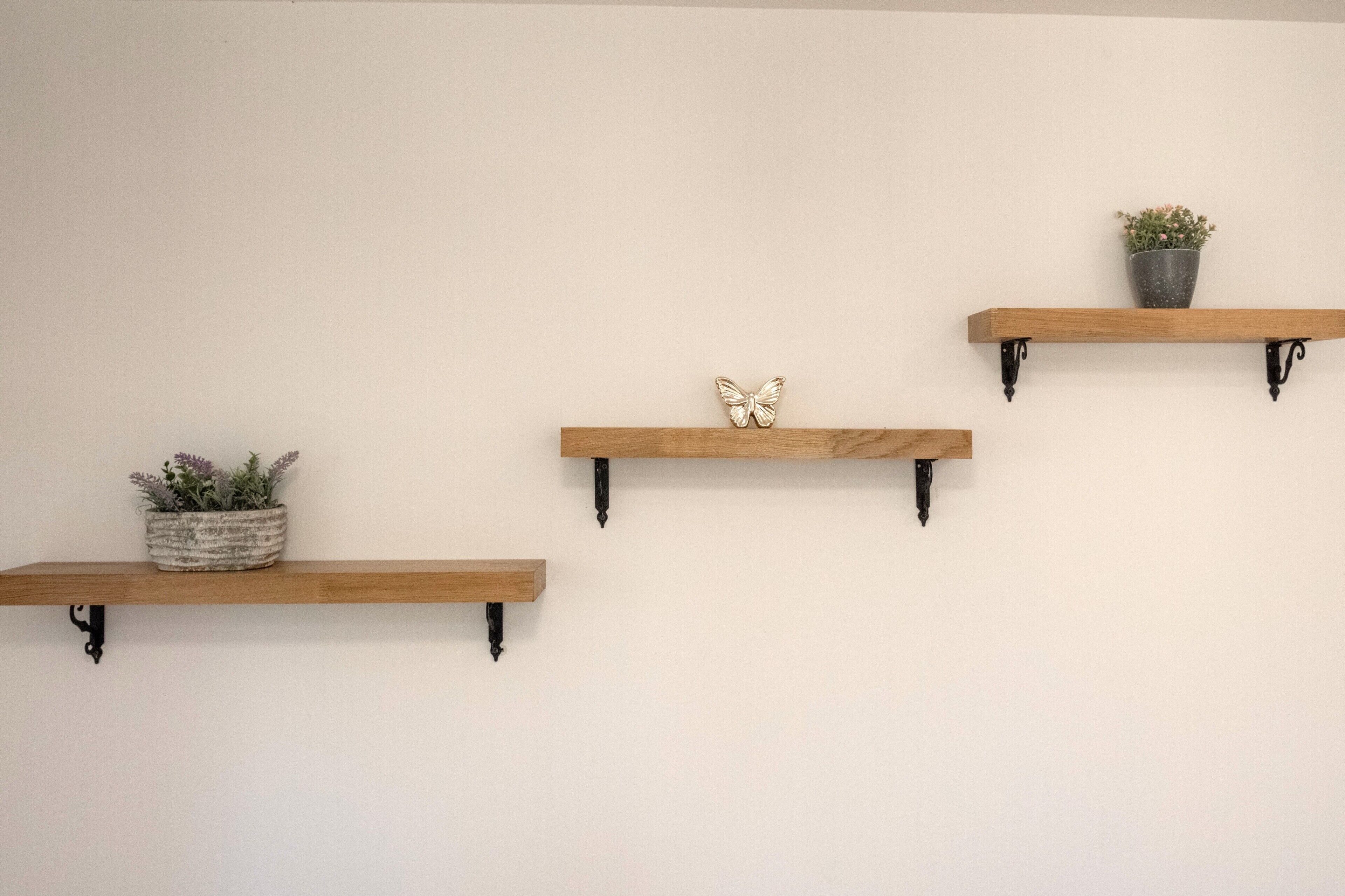 Trois étagères en bois, soutenues par des équerres noires, sont fixées à un mur uni. Chaque étagère porte un pot de plante et un petit objet décoratif.