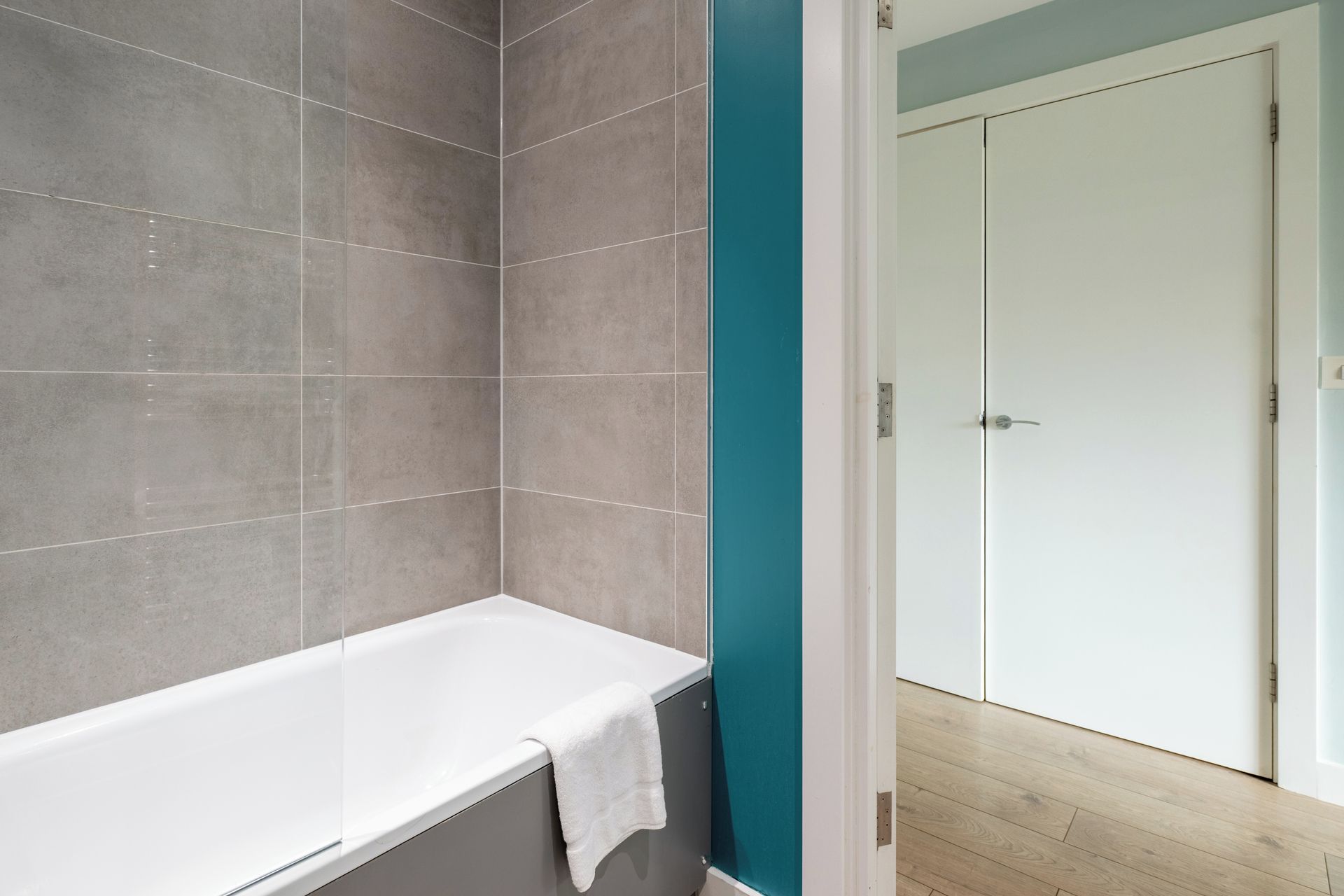 Ein Badezimmer mit Badewanne, grauen Fliesen, einer türkisfarbenen Akzentwand und einer weißen Tür.
