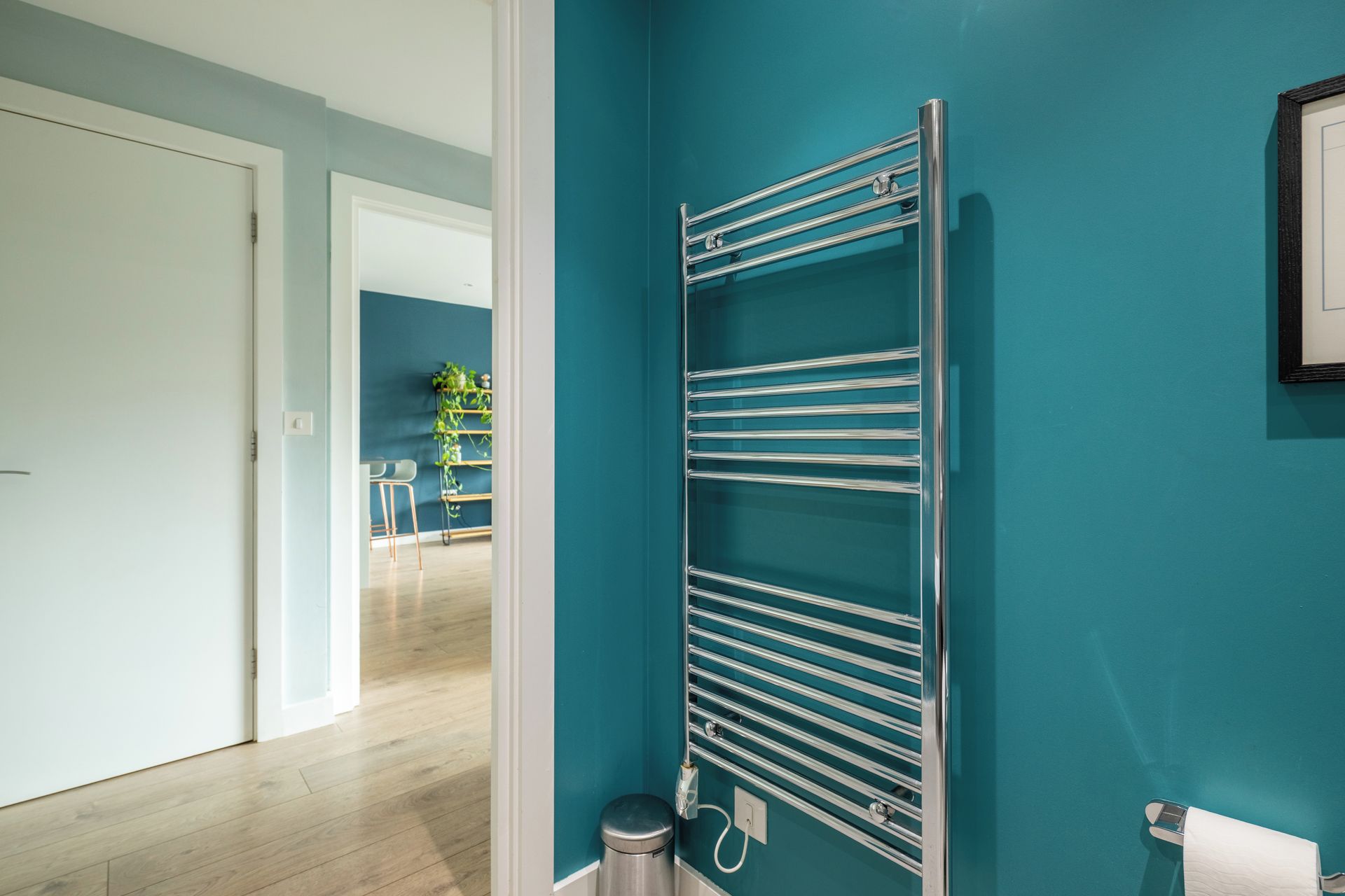 Un pasillo con pared verde azulado, un perchero metálico y una puerta que da a otra habitación.